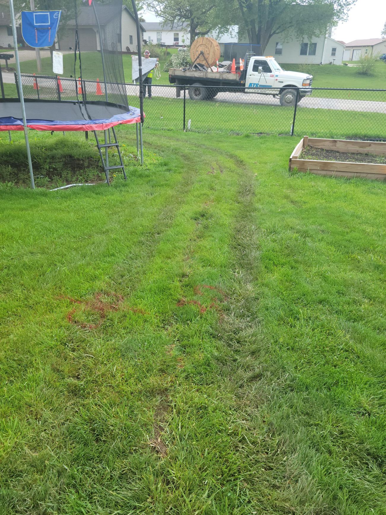 Tire tracks in green grass lead to a truck with equipment in a backyard next to a trampoline and garden bed.