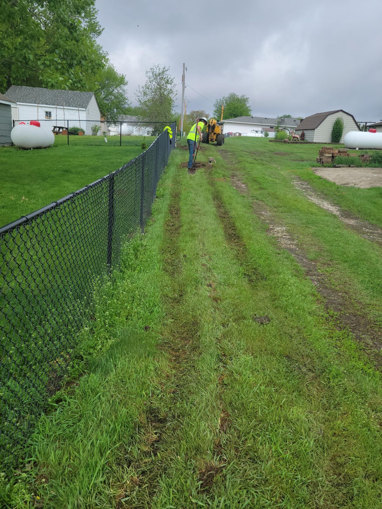 Workers near a black chain link fence and a tractor on a grassy path. Overcast day.