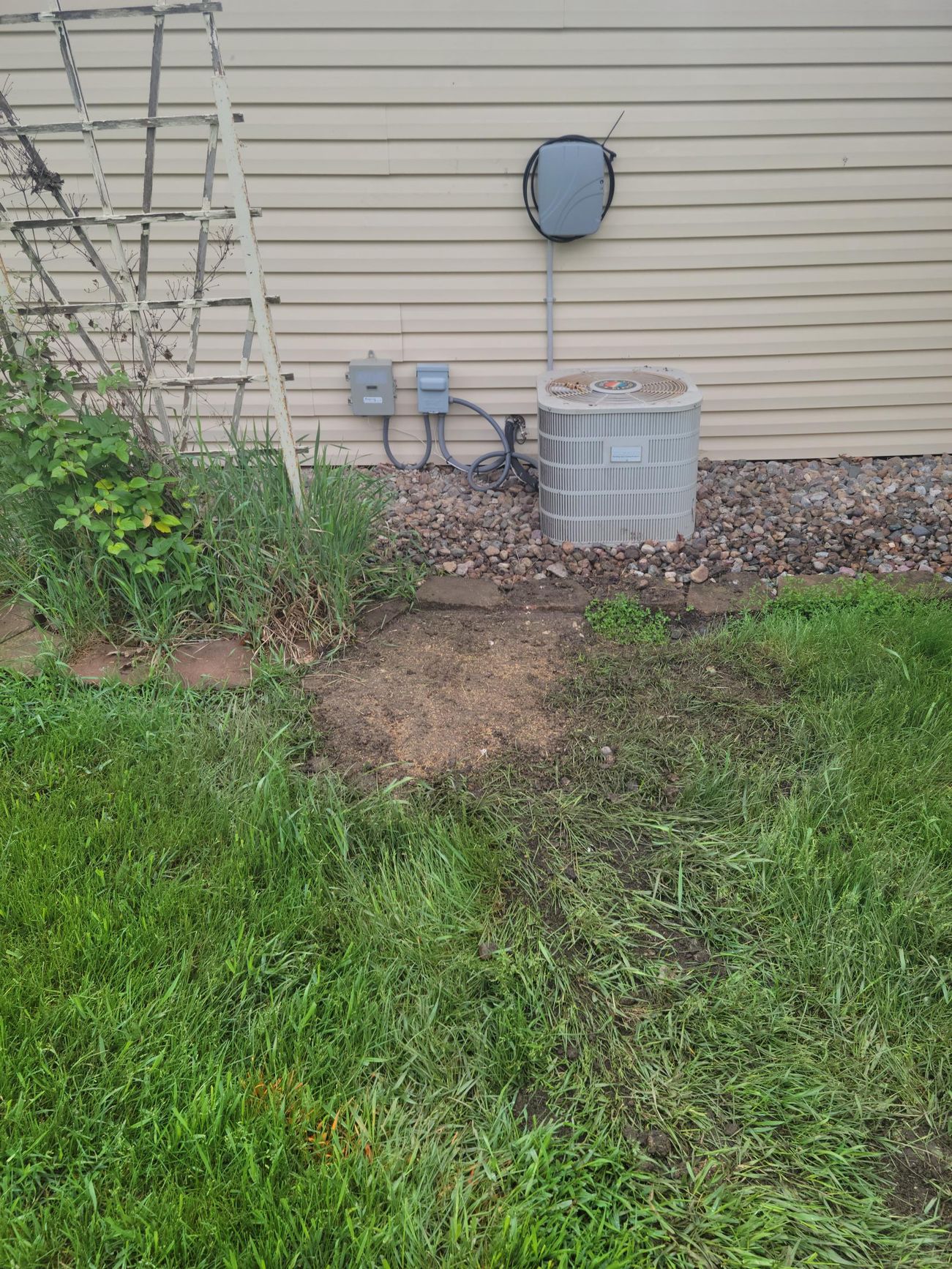 AC unit next to a house with electrical boxes and a trellis in a grassy yard.