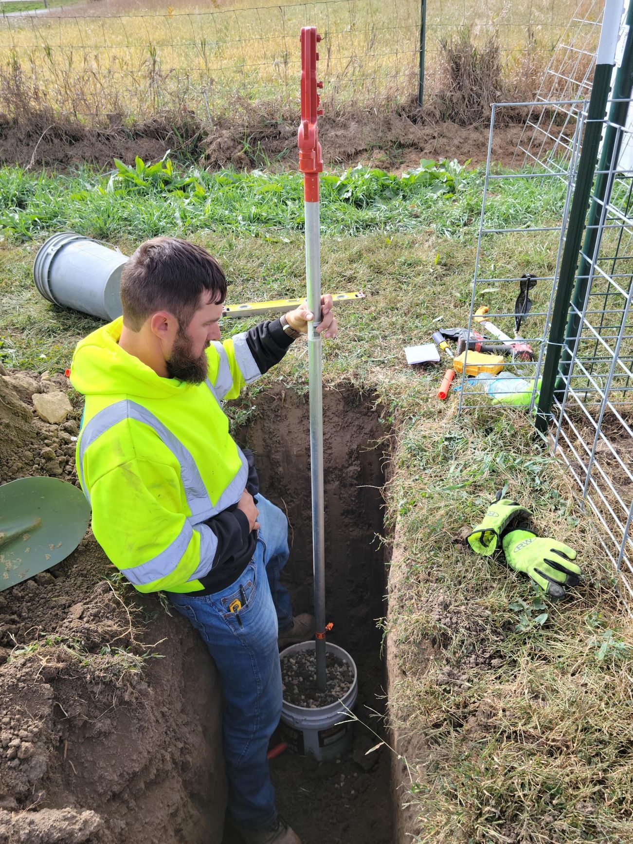 Person in high-vis vest in a trench, using a tool to work on an underground object near a fence.
