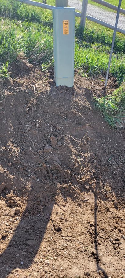 A rectangular utility box in the dirt near a fence. A hazard warning is on the box.
