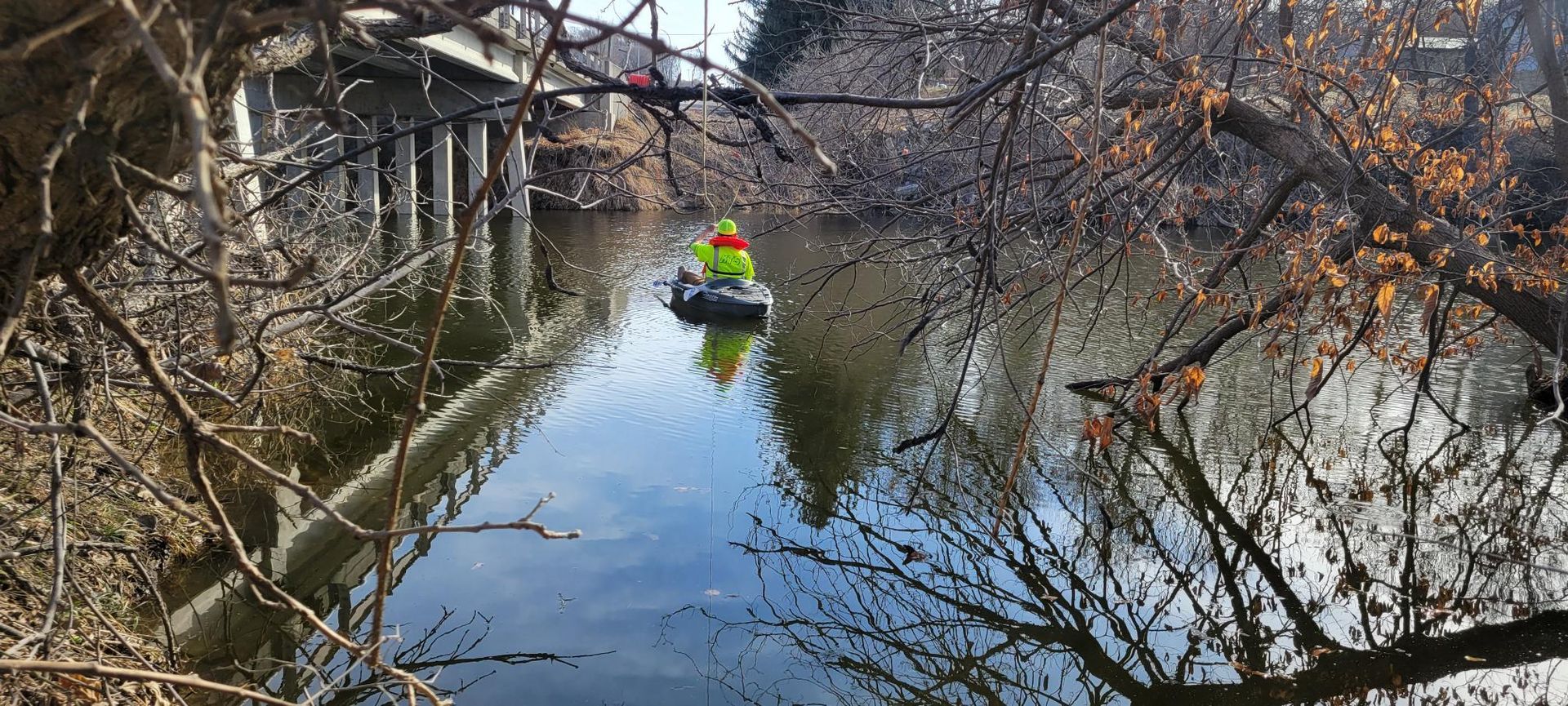 Person in yellow work clothing on a small boat in a dark body of water, under a bridge and surrounded by bare trees.