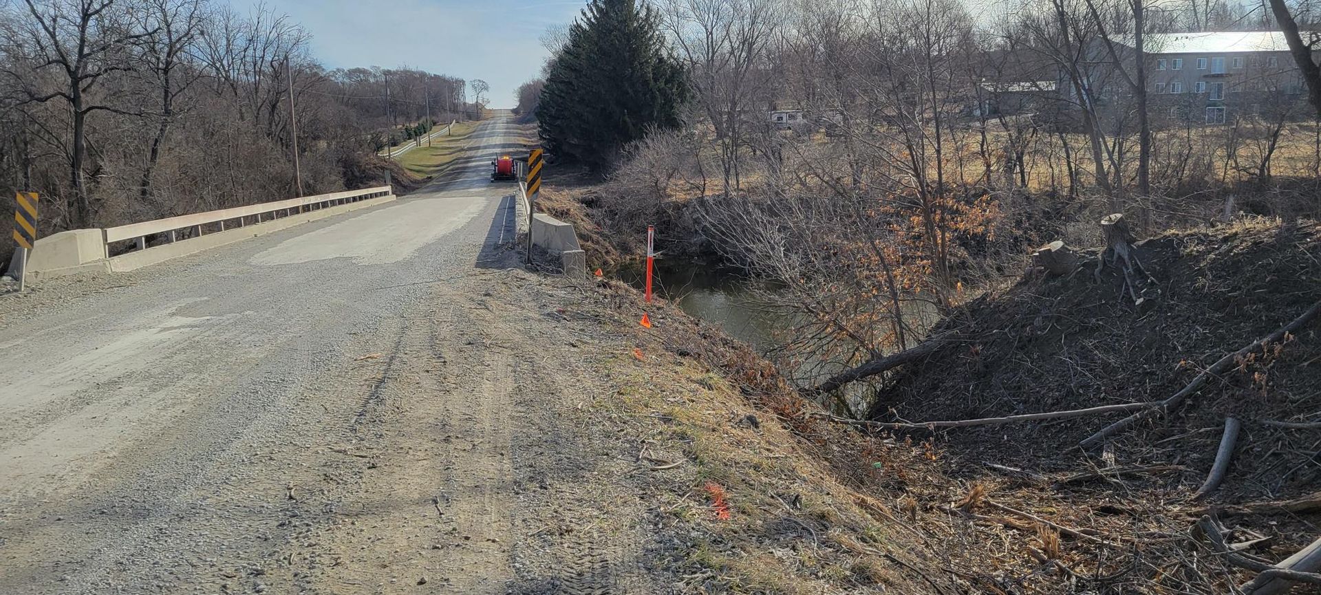Gravel road over a bridge in a rural area. A red car is driving toward the bridge. Bare trees line the road.
