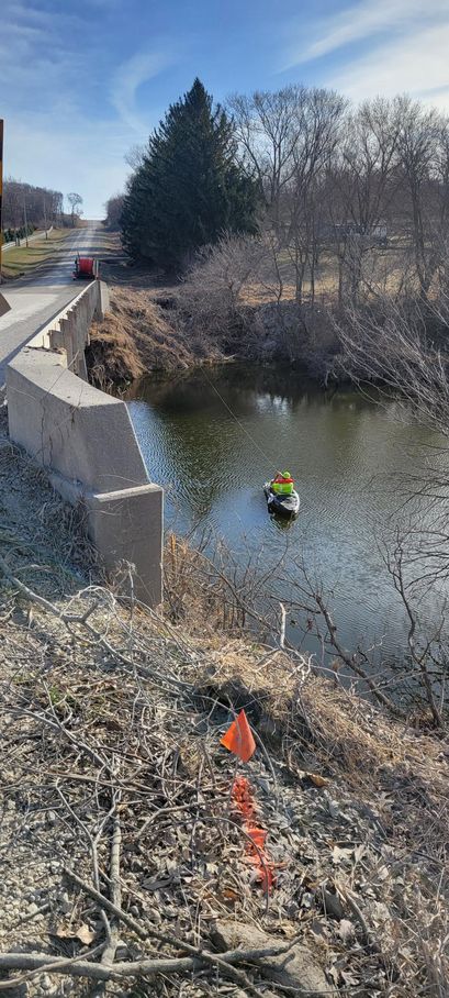 A boat with a person in a yellow vest in a river by a bridge. Dry vegetation and a road are visible.