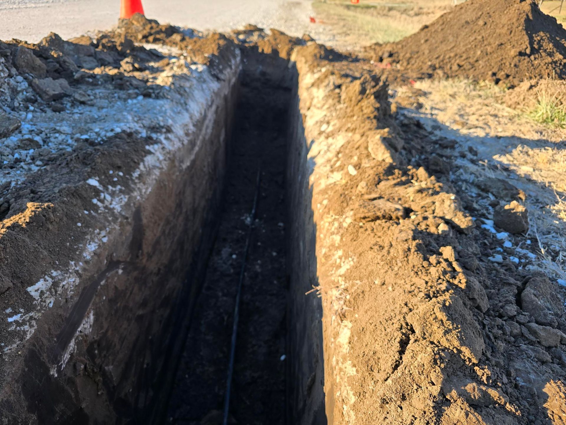 Long, deep trench dug in dirt, likely for burying utilities, with road and orange cone visible.
