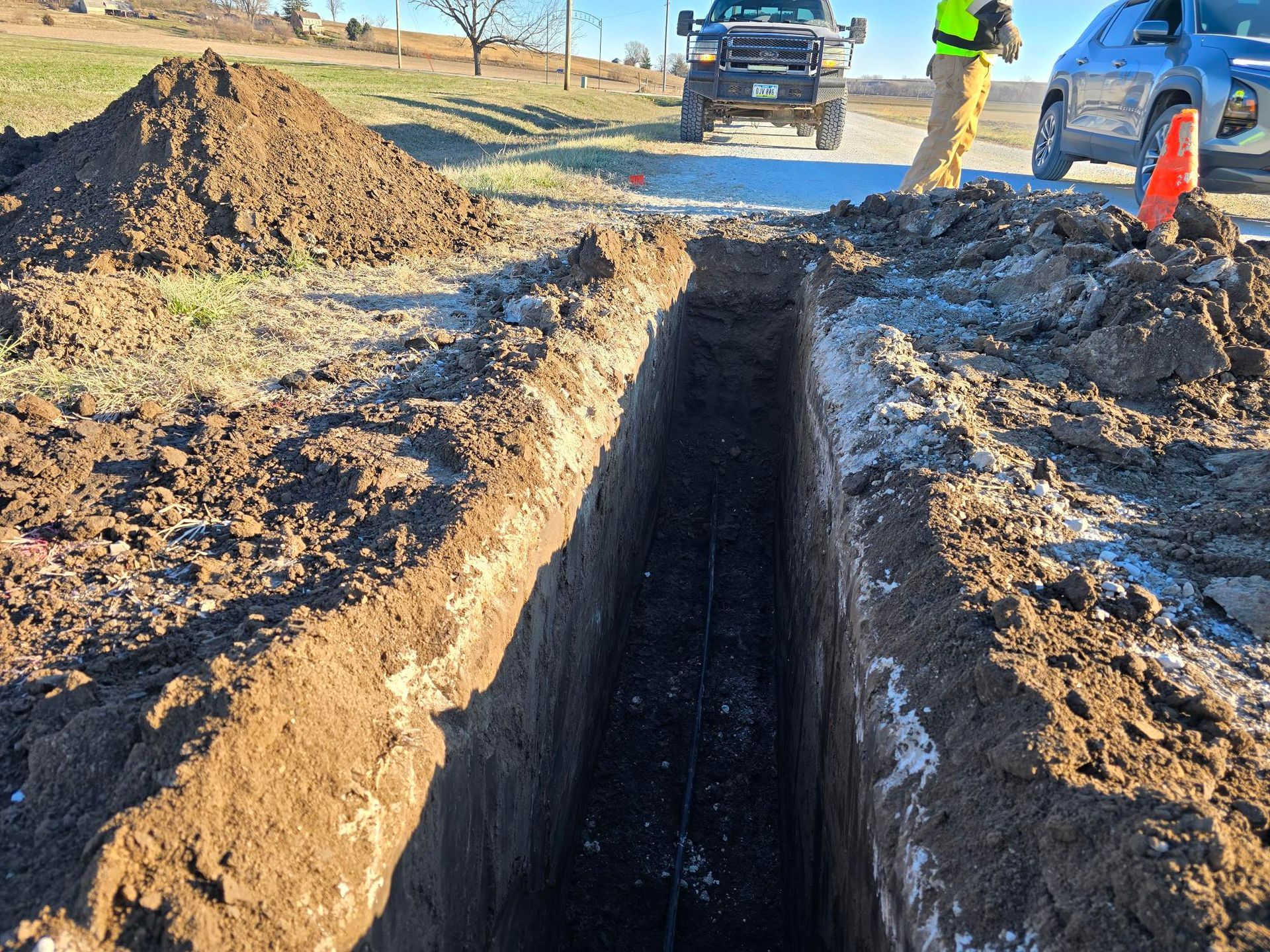 Long trench dug in dirt, likely for utility work, with trucks and worker in background.