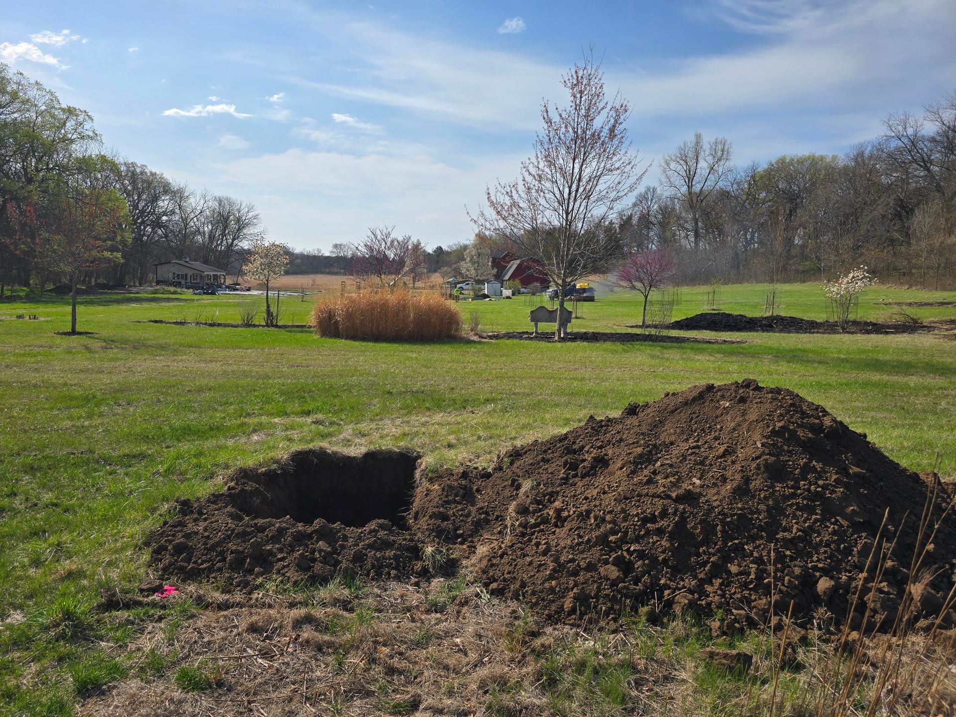 Hole dug in a grassy yard, with a mound of dirt beside it. Trees and a sunny sky in the background.