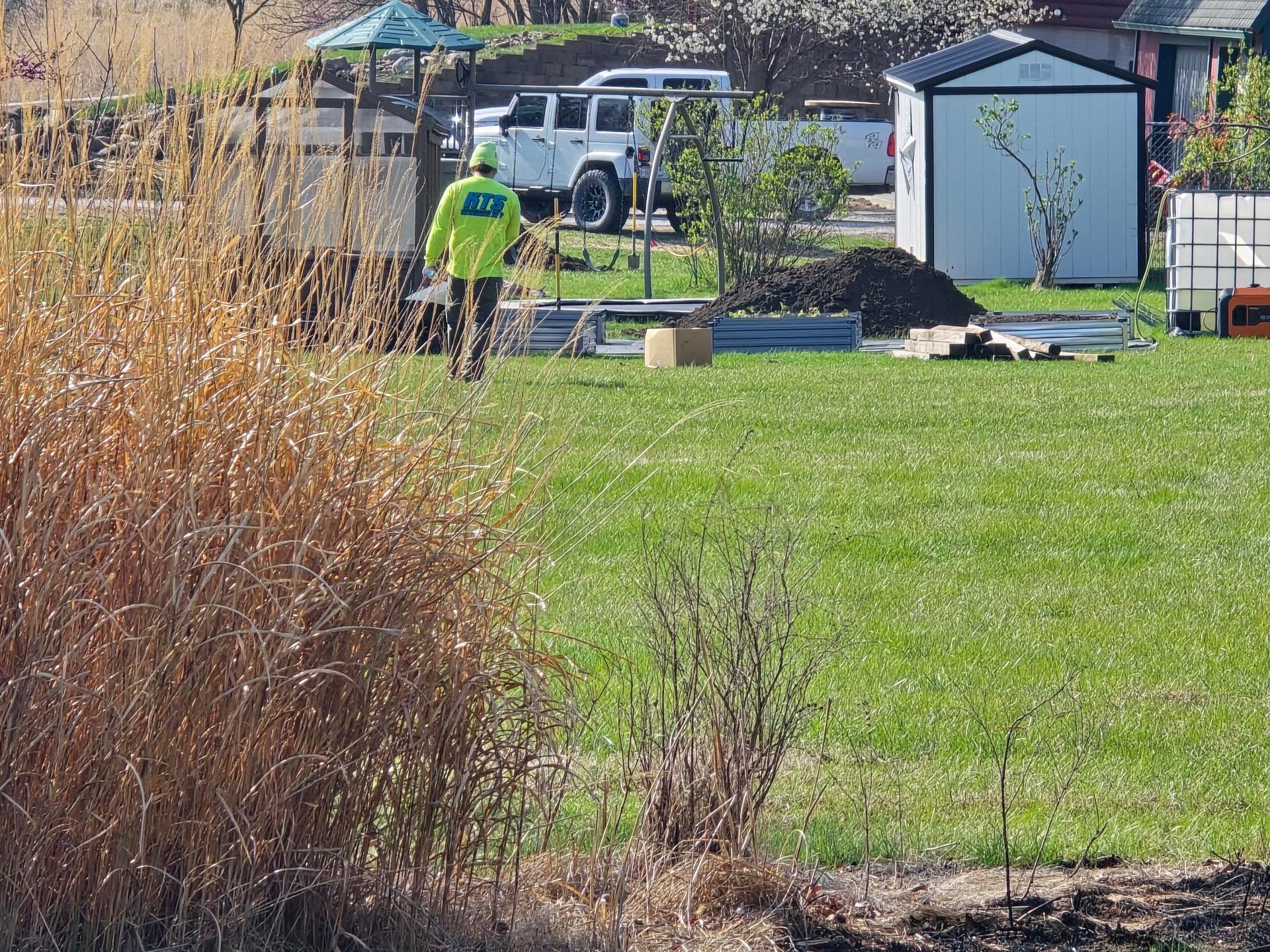 Man in neon vest stands on green grass near a shed and vehicles. Dried brown plants in foreground.