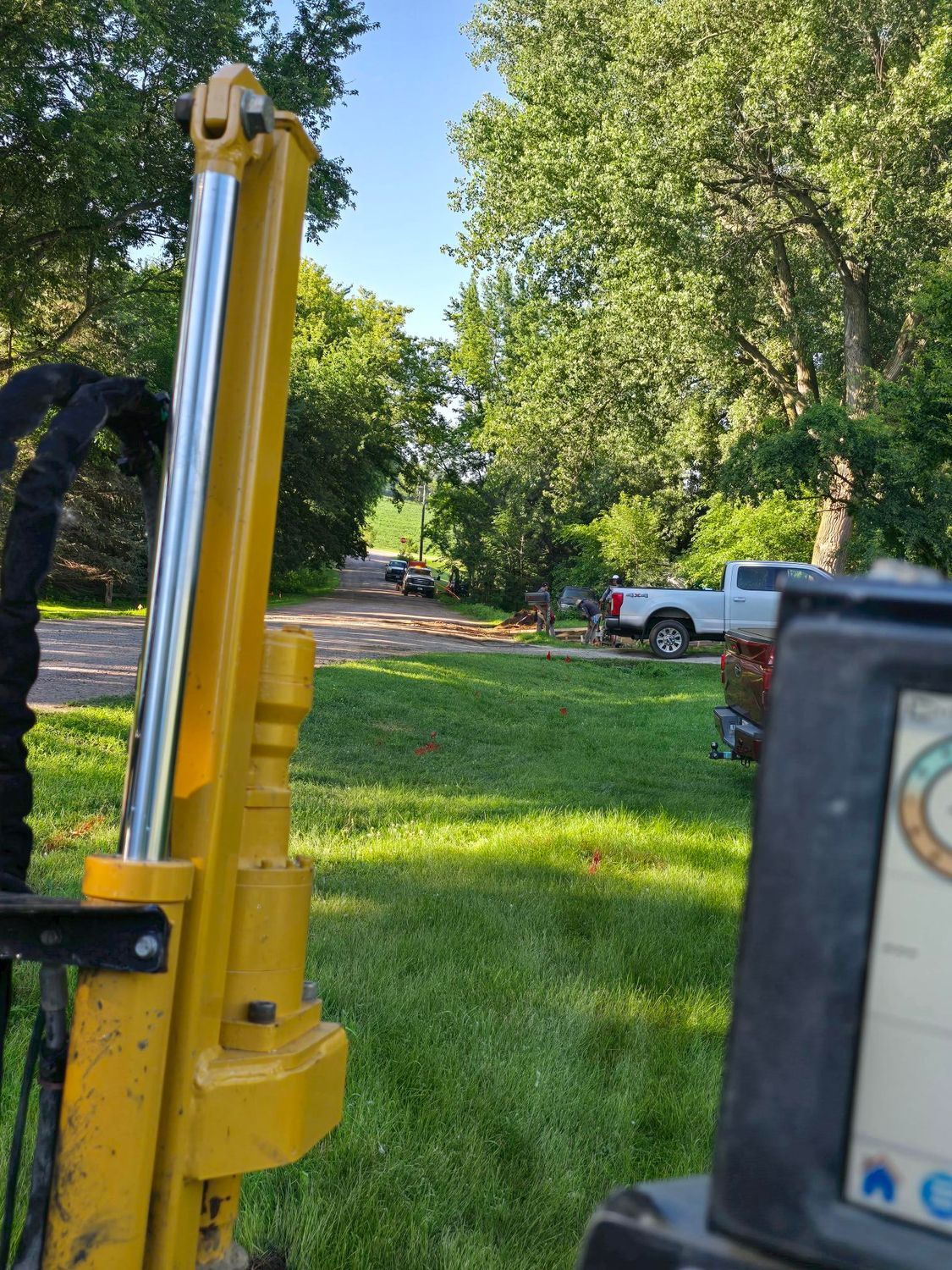 Yellow hydraulic arm, display screen, view of grassy yard, road, and parked trucks on a sunny day.