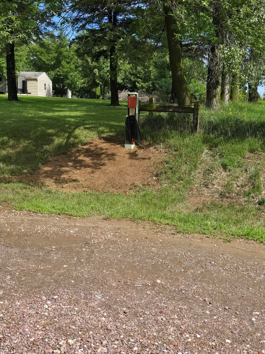 Gravel road leads to grassy area with mailbox, trees, and small building in the background.