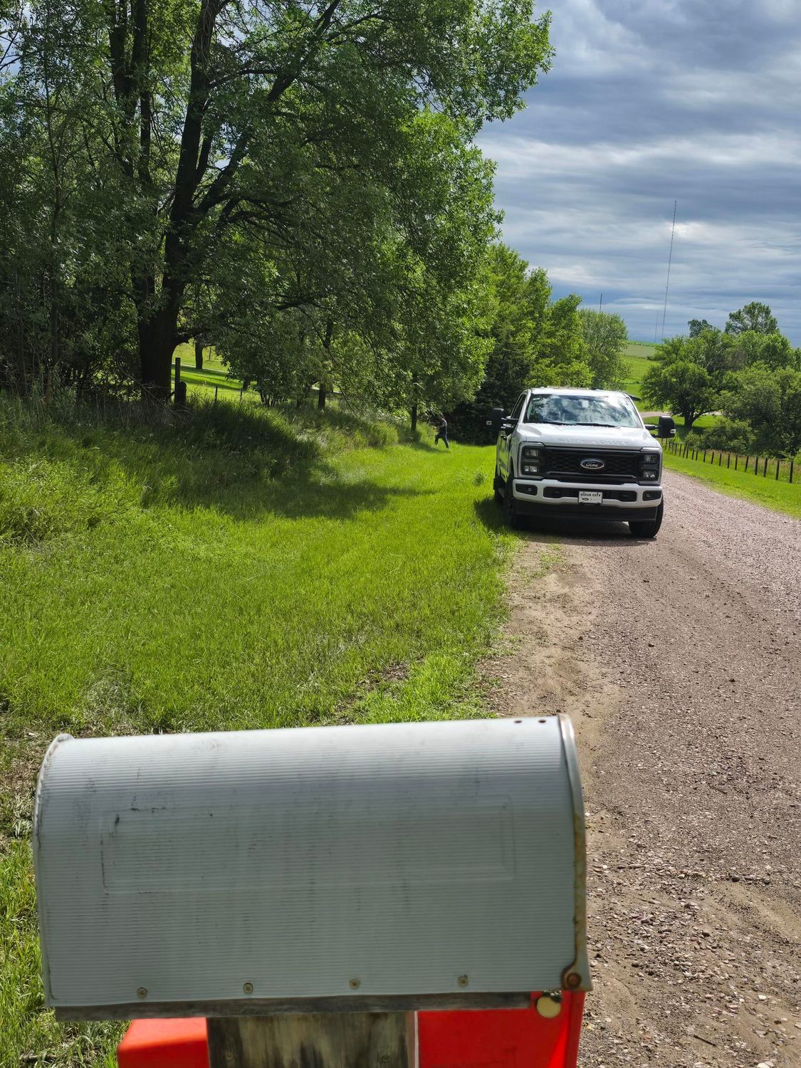 Mailbox in focus, a white truck on a gravel road, and green trees under a cloudy sky.