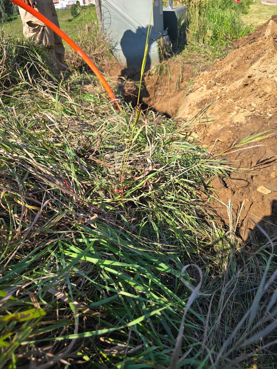 A trench in soil with orange tubing entering a gray utility box, next to overgrown grass.