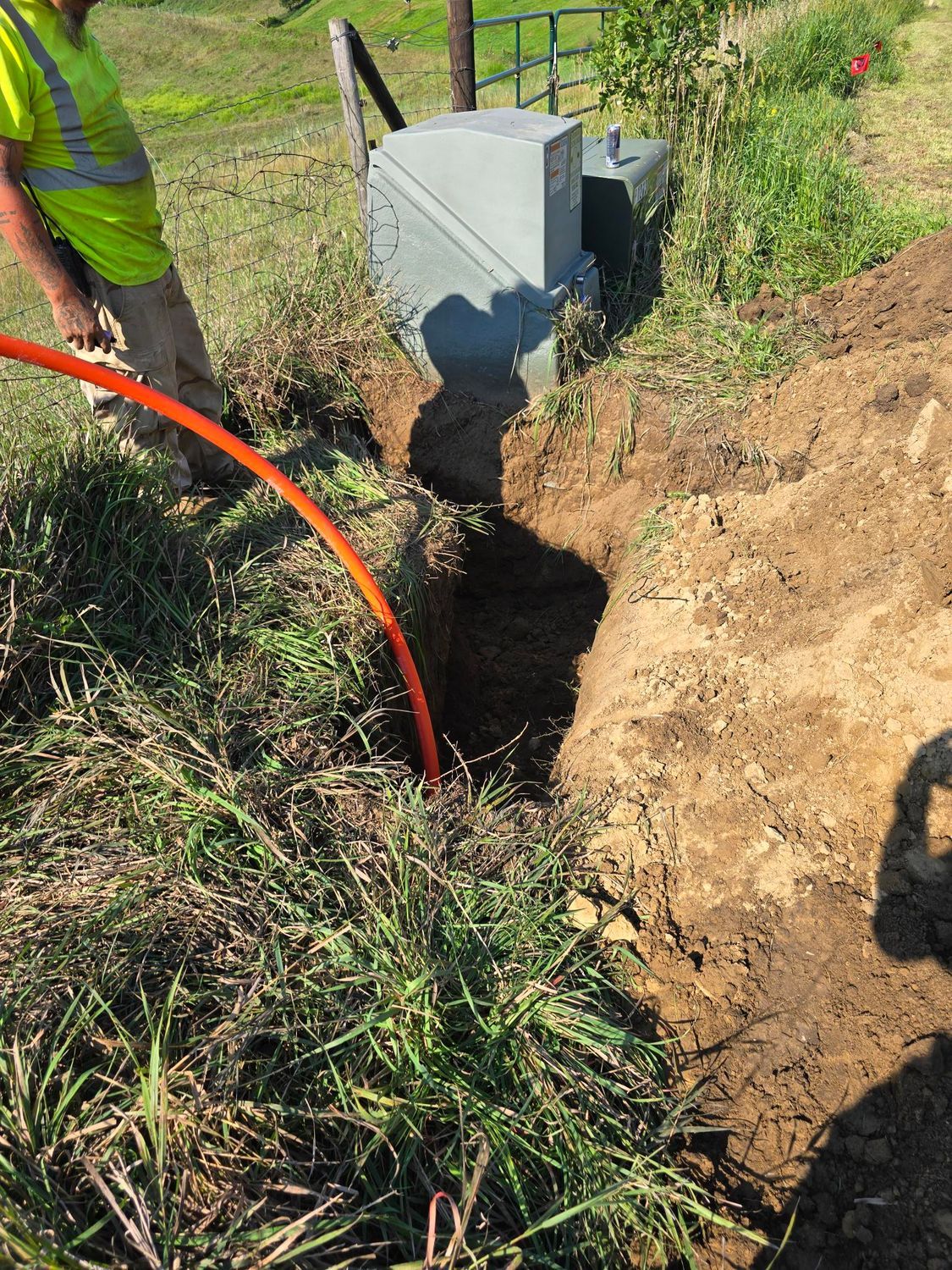 Man in safety vest installing orange cable near metal utility box in grassy field.