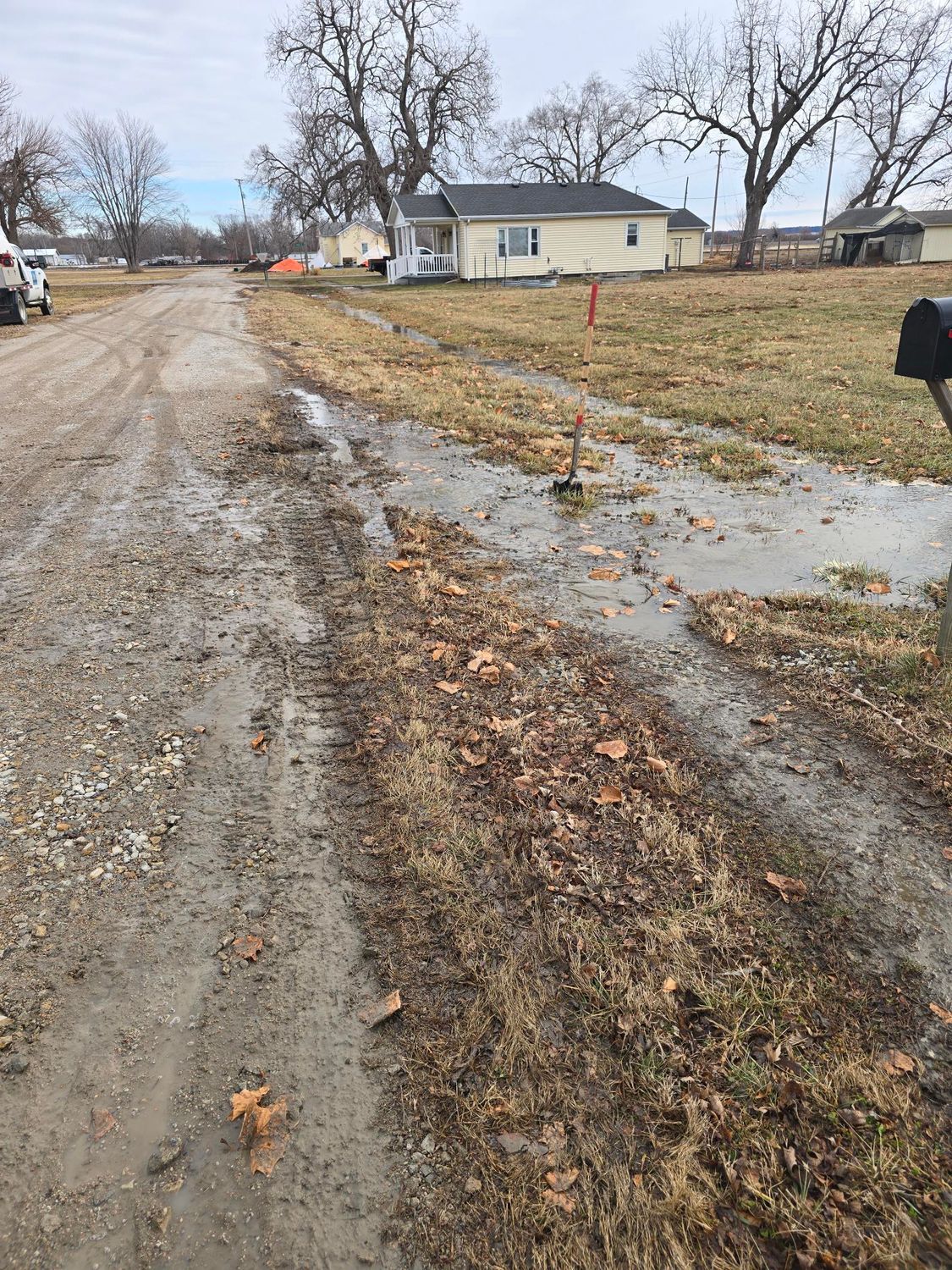Muddy dirt road with standing water, small house in background. Overcast, outdoor scene.