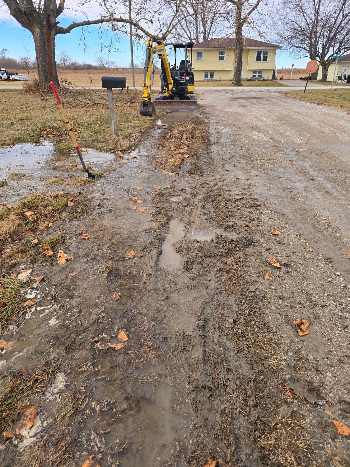 A muddy driveway with an excavator, leaves, and standing water in front of a house.