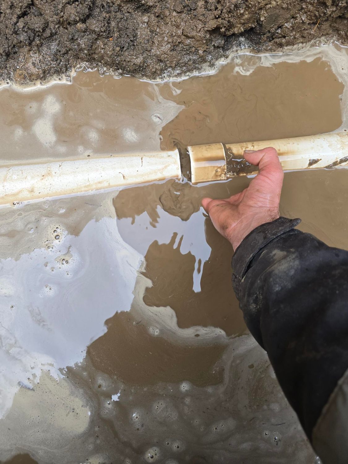 Hand holding broken white pipe in muddy water.