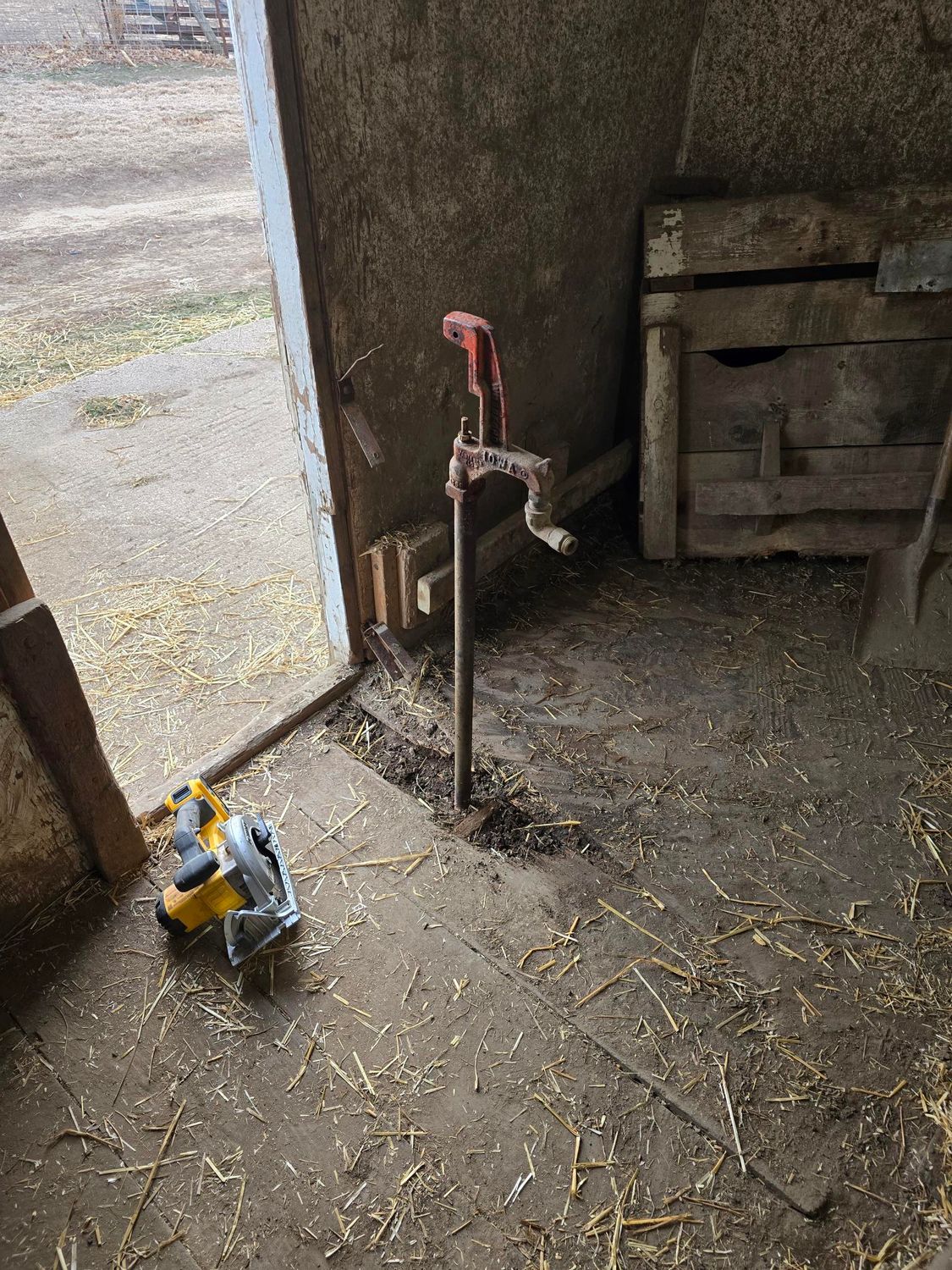 Inside a barn, a metal water spigot stands, next to a power saw, and a wooden door.