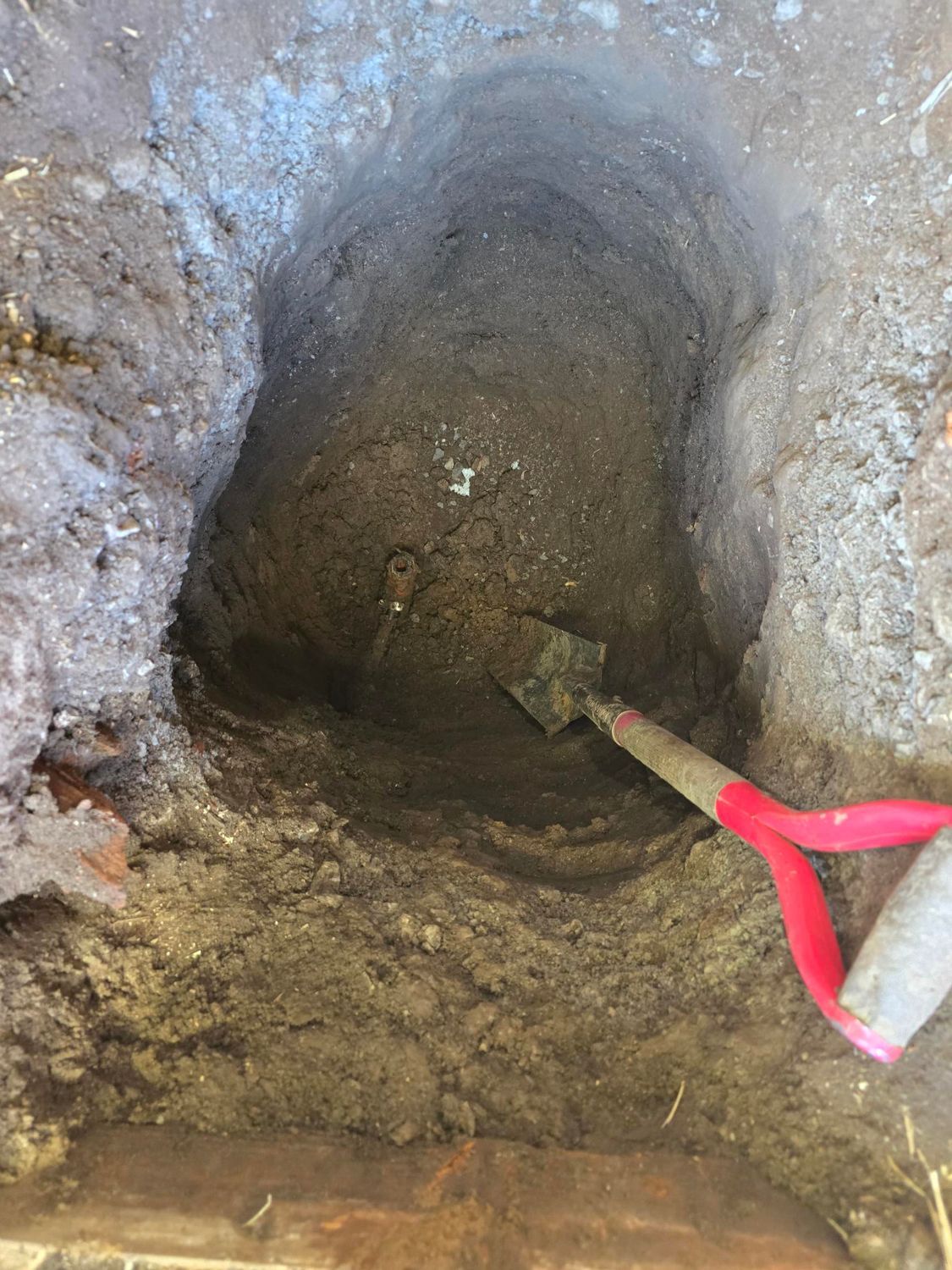 Hole dug in the ground with a red-handled shovel resting inside. Earth and brick surround.