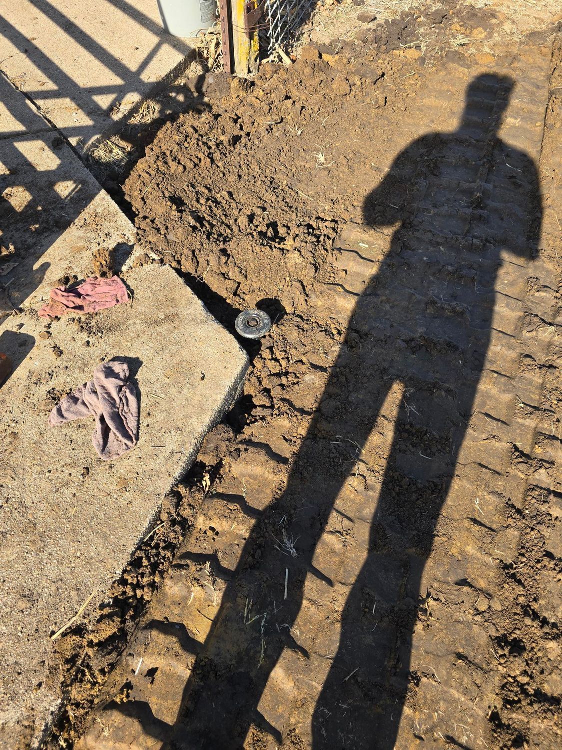 Shadow of person standing near excavation in dirt, with concrete and work gloves.