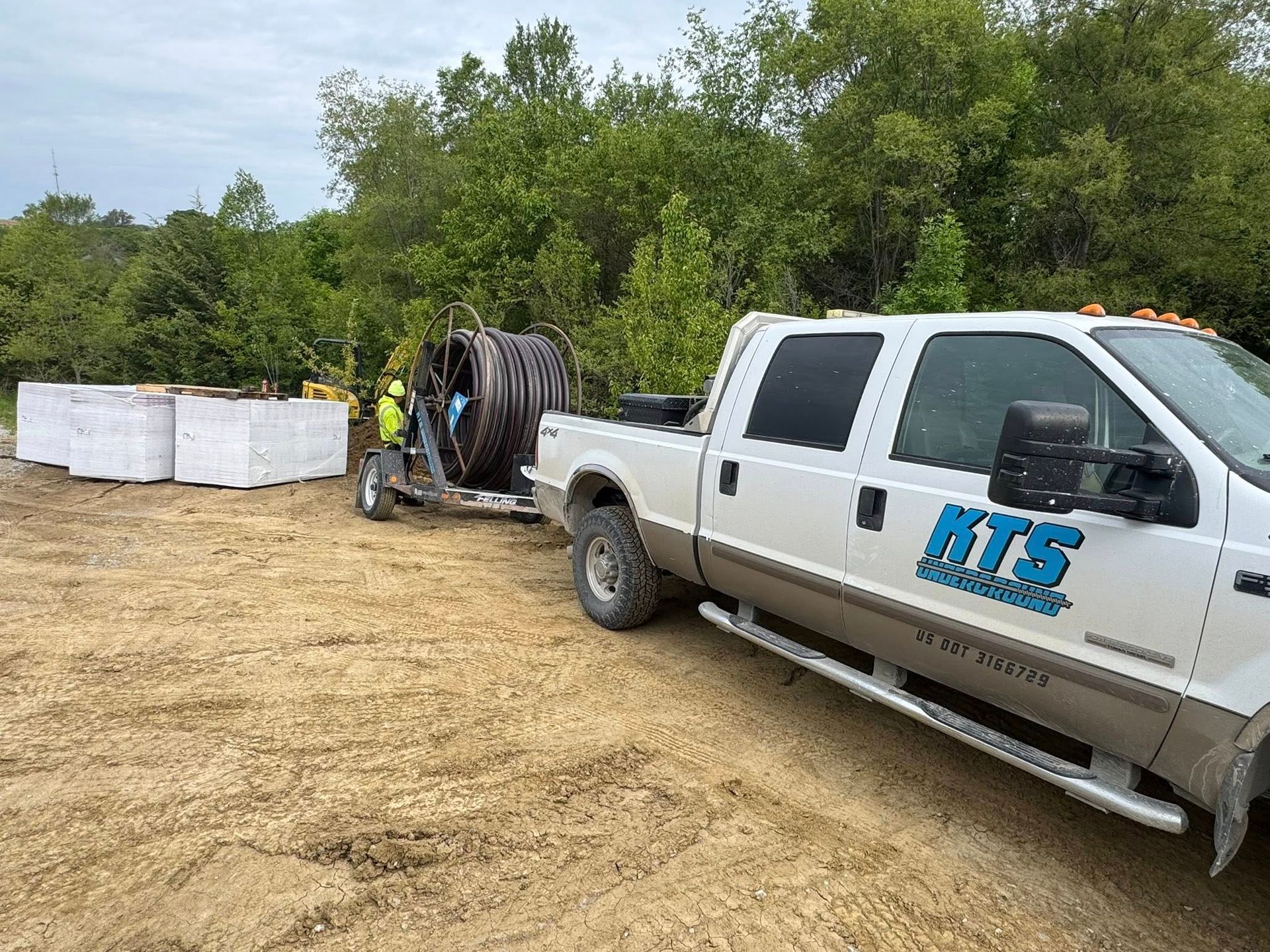 White truck towing a cable reel; worker near it on a dirt road, alongside concrete blocks and foliage.