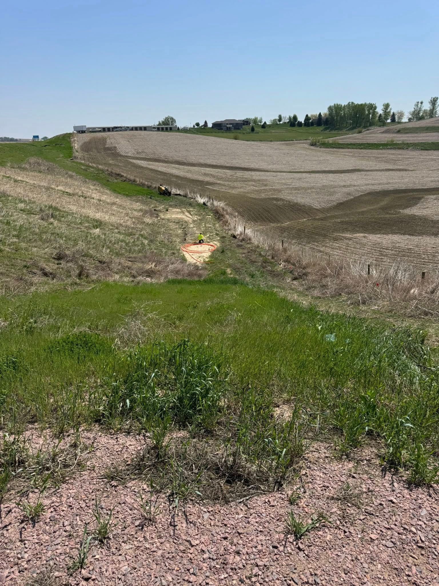 Grassy area with gravel, leading into a dry, barren, dirt field. Blue sky, distant houses.