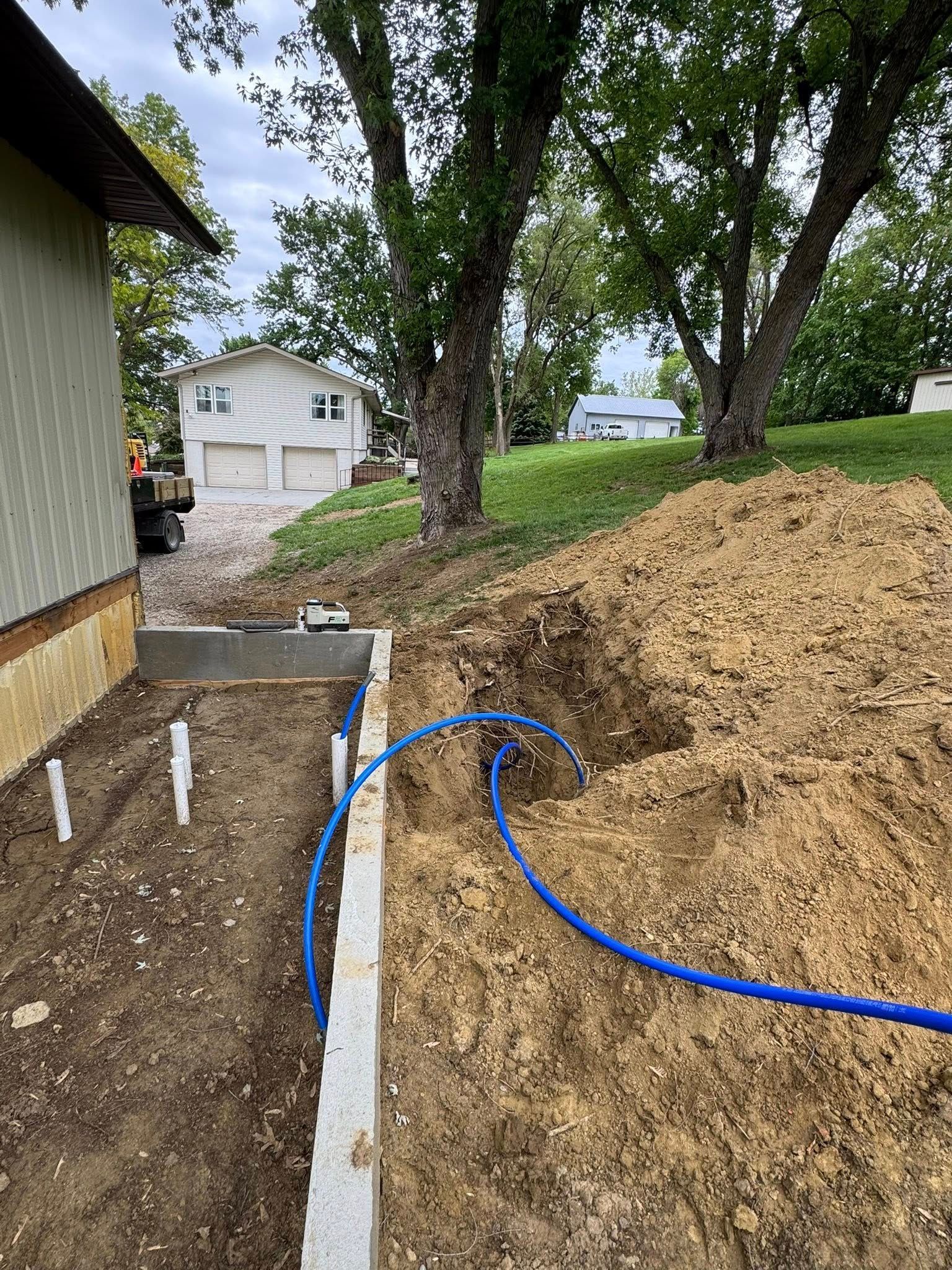 Construction site: blue pipes in a trench near a house foundation and dirt pile. Trees and other houses in background.