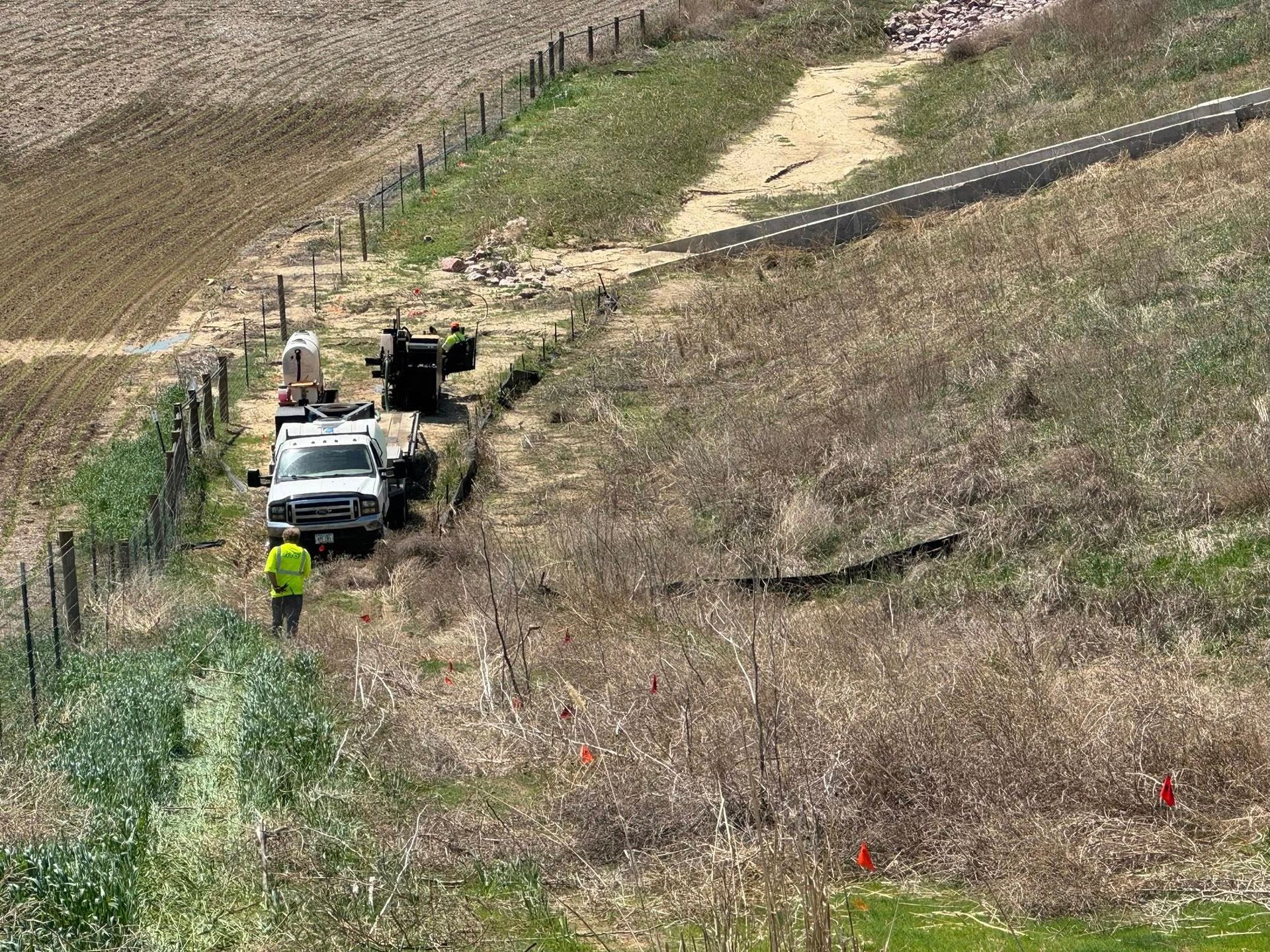 Trucks and a worker on a hillside, possibly for road or utility work. Fencing and road visible.