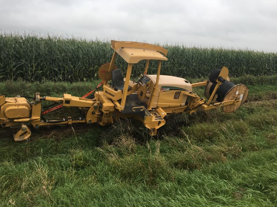 Yellow trenching machine in a field of grass next to a cornfield, cloudy sky.