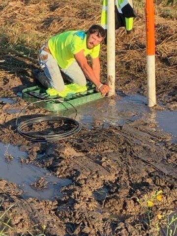 Man in neon shirt working near a white post in muddy ground. A green utility box is nearby.