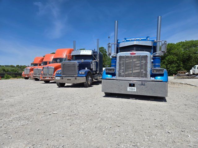 Line of semi-trucks, blue and orange, parked on gravel in bright sunlight.