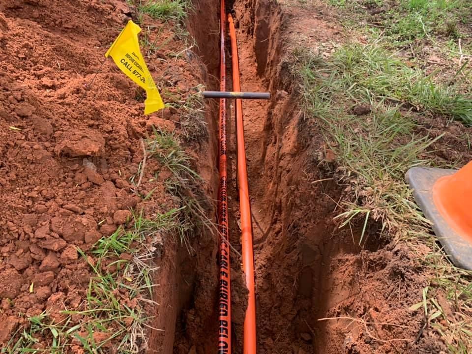 A trench dug in the ground with orange utility lines and a caution tape. A yellow 