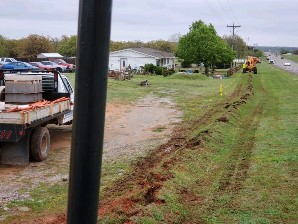 A work truck and tractor on a roadside. A trench is dug through the grass. Houses are in the background.