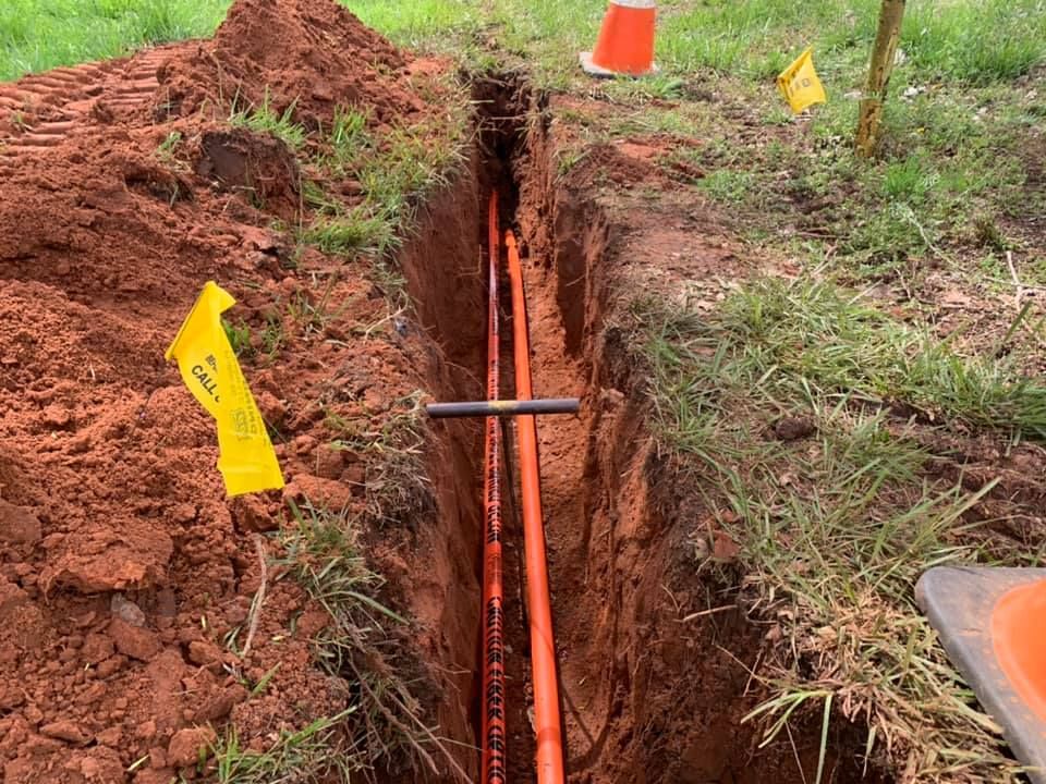 A trench dug in red soil, containing two orange cables, marked with flags and a traffic cone.