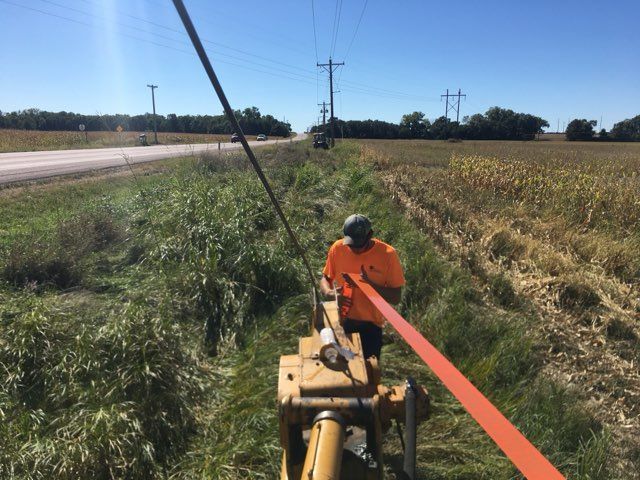 Worker in orange vest pulling an orange cable from a machine in a ditch beside a road.