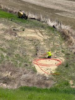 Person in yellow vest working with orange tubing on a hillside. A yellow vehicle is visible in the background.