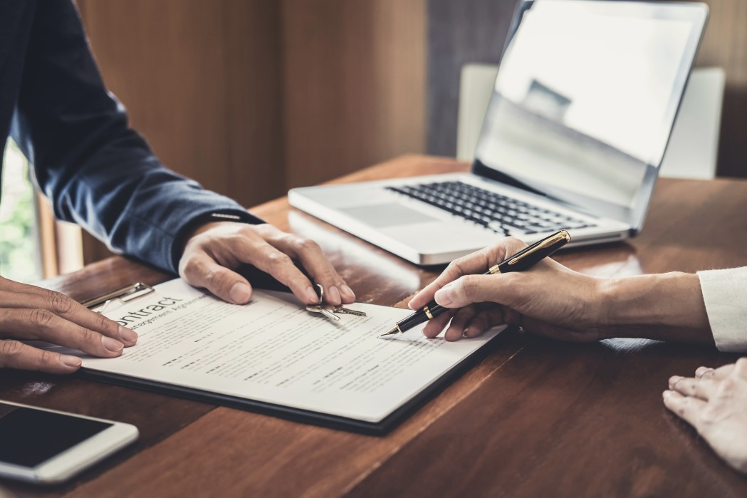 Person signing a document with a pen, laptop and phone on a wooden table.