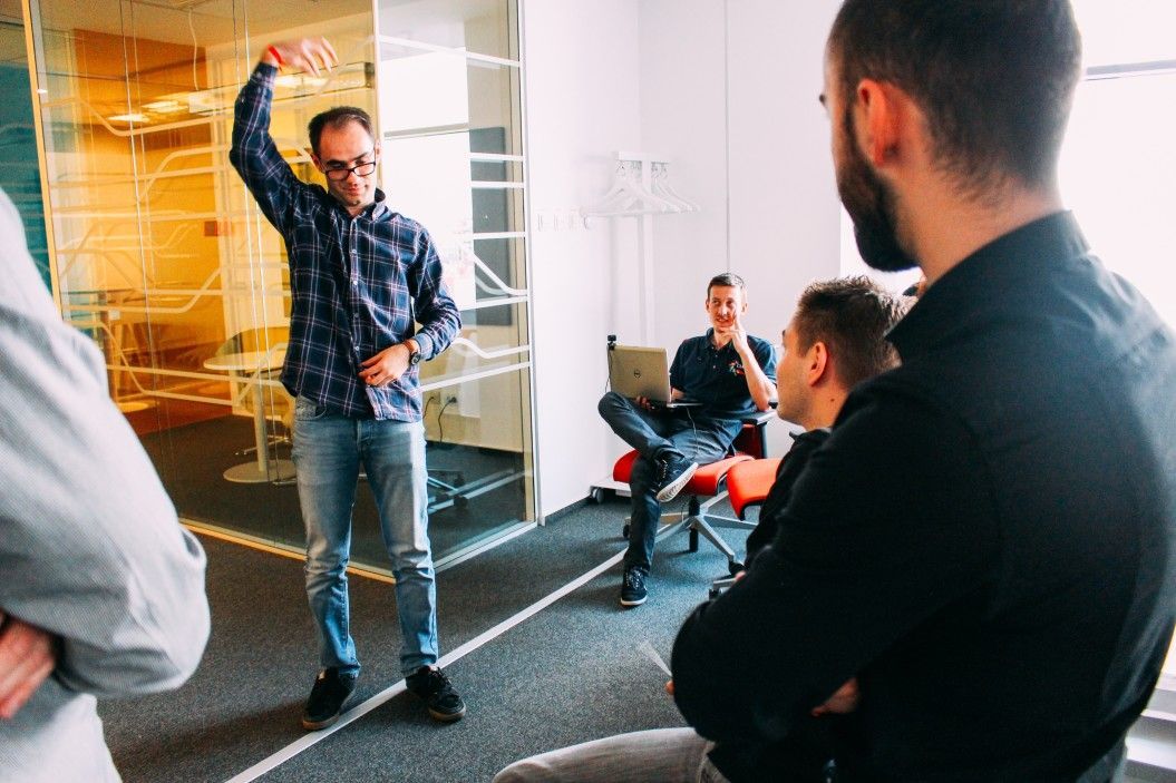 Man in blue shirt gestures in meeting, facing other men. One man uses a laptop. Modern office.