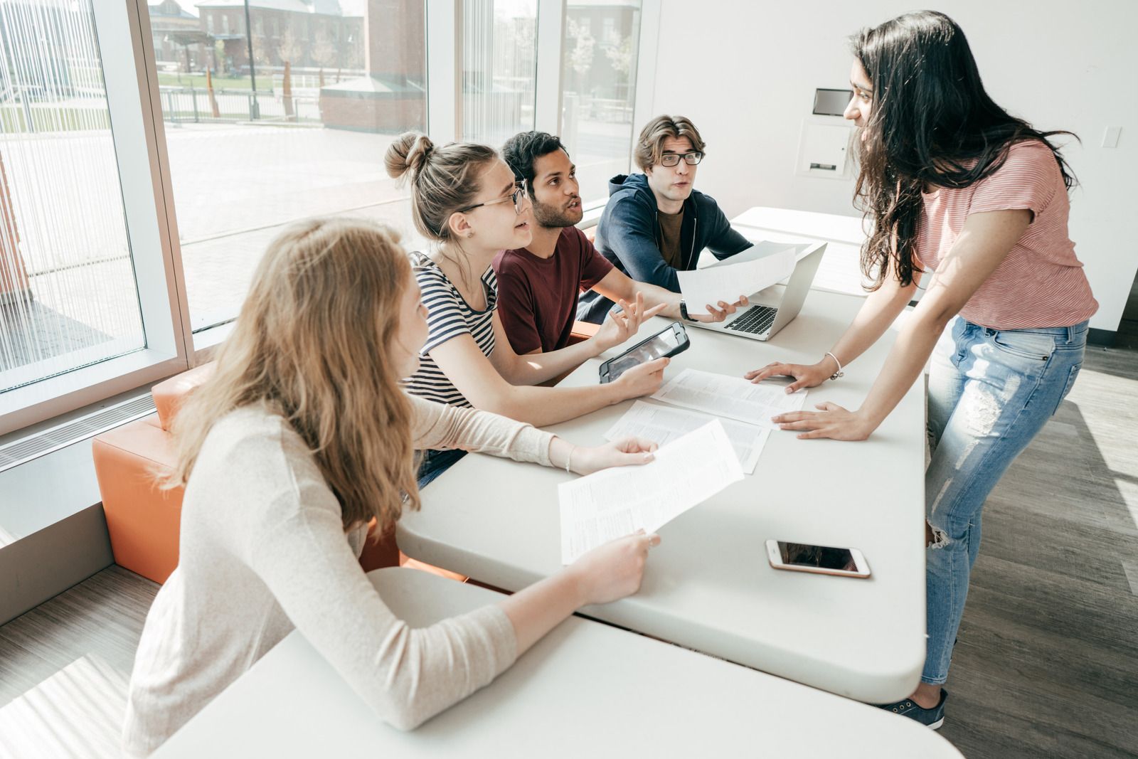 Five people collaborate at a table, reviewing documents, laptops and tablets in bright room.