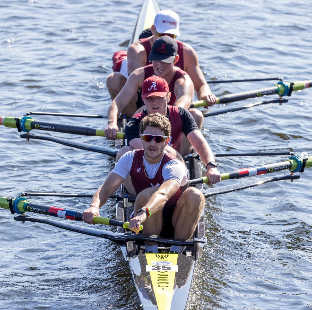 Rowing team in a boat on water, wearing maroon and white. Rowers are focused, pulling oars with effort.