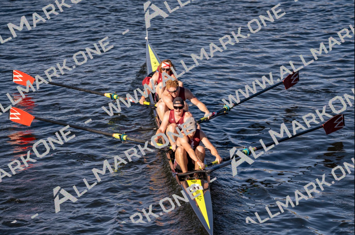 Rowing team in a boat, moving across water. The team wears red and black, with oars dipping in the water.