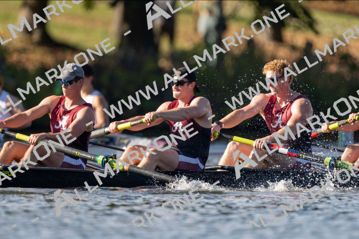 Rowers in a dark rowing shell on the water, wearing maroon and white. They are pulling oars, with splashes.