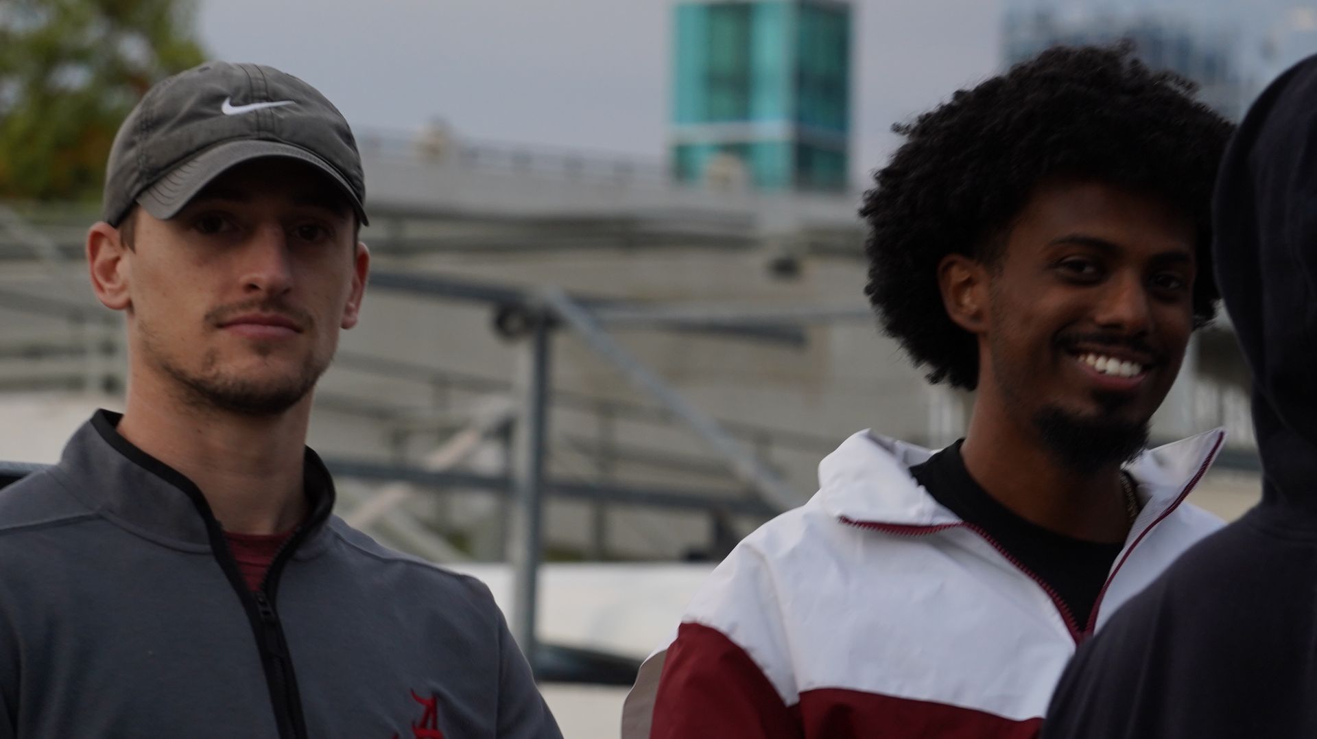 Two men smiling outdoors; one in a cap and jacket, the other with curly hair and a white and maroon jacket.