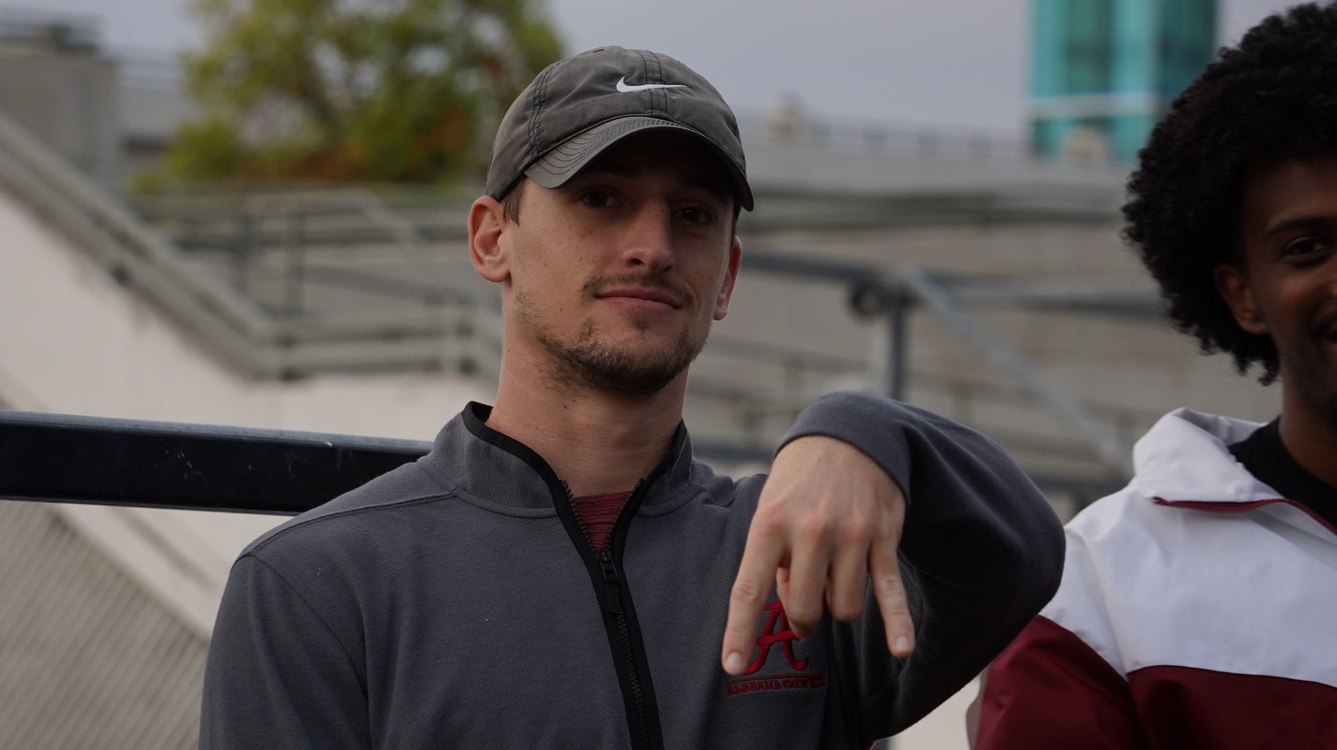 Man in gray jacket and cap pointing down, smiling, with another man partially visible on the right.