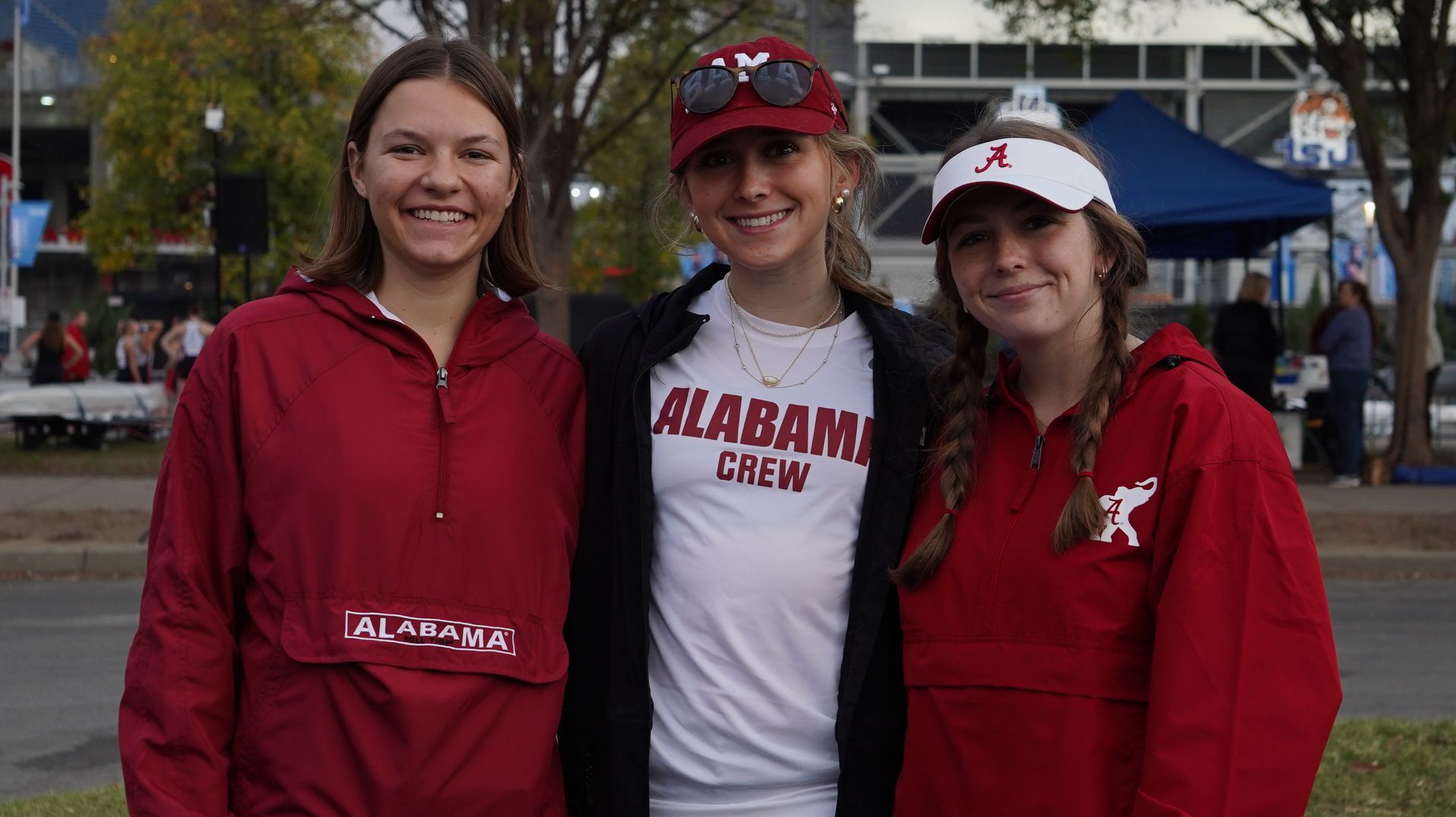 Three young women smiling for a photo, wearing Alabama Crimson Tide gear outdoors.