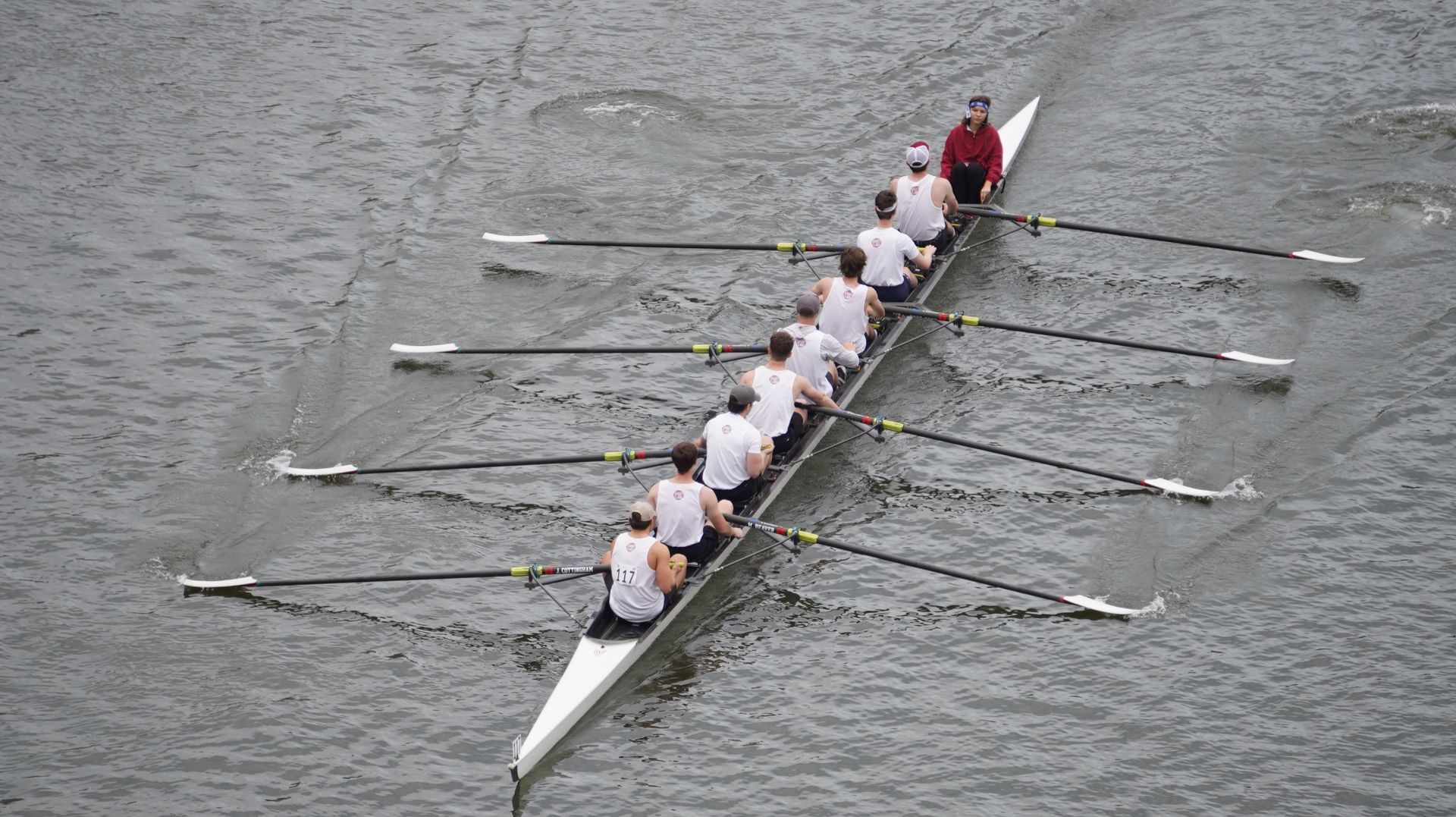An overhead shot of a crew team rowing in a long, white boat on water. Eight rowers and a coxswain are visible.