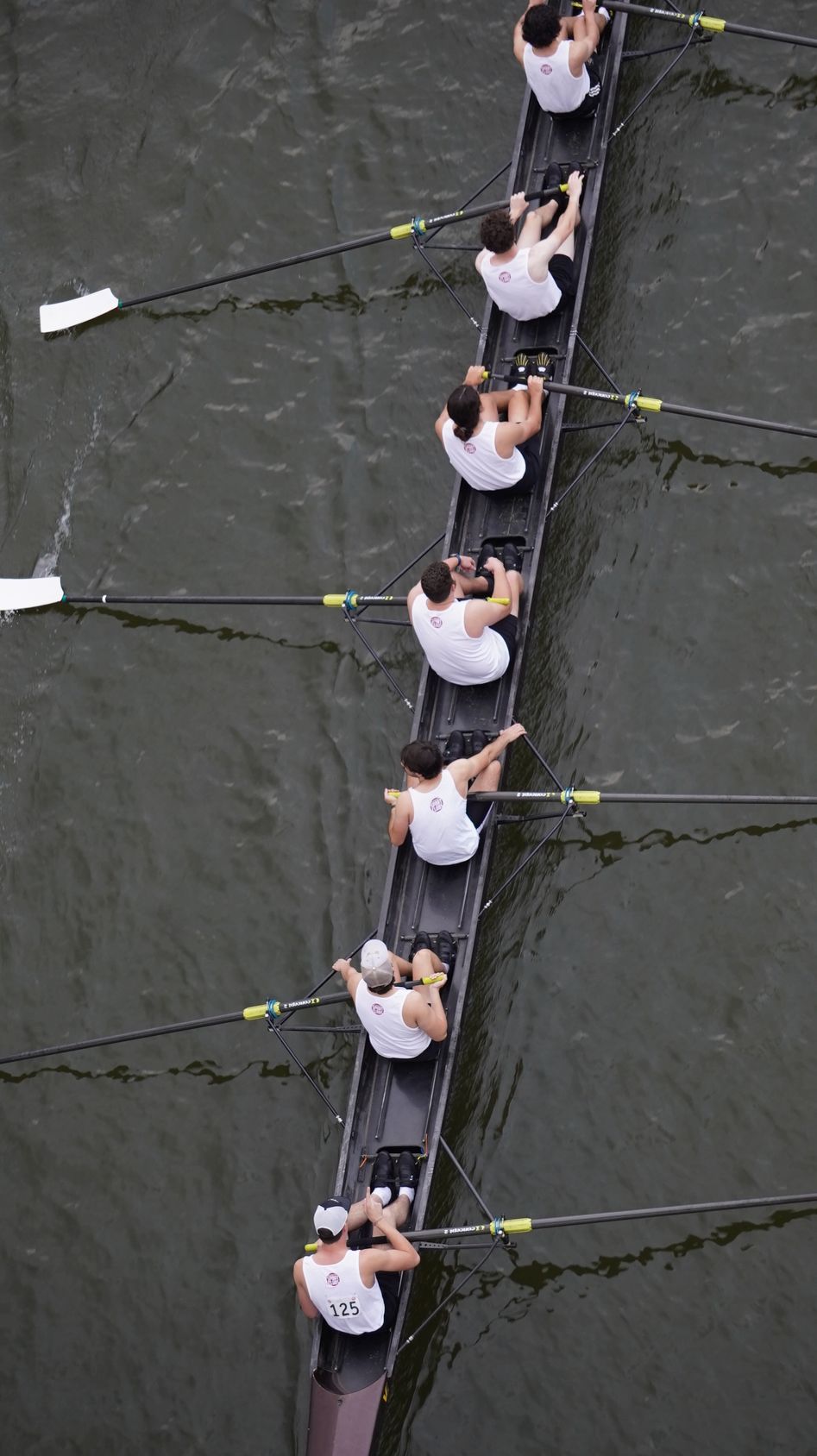 Overhead view of a rowing team in a dark-hulled boat on water, all rowing in unison.