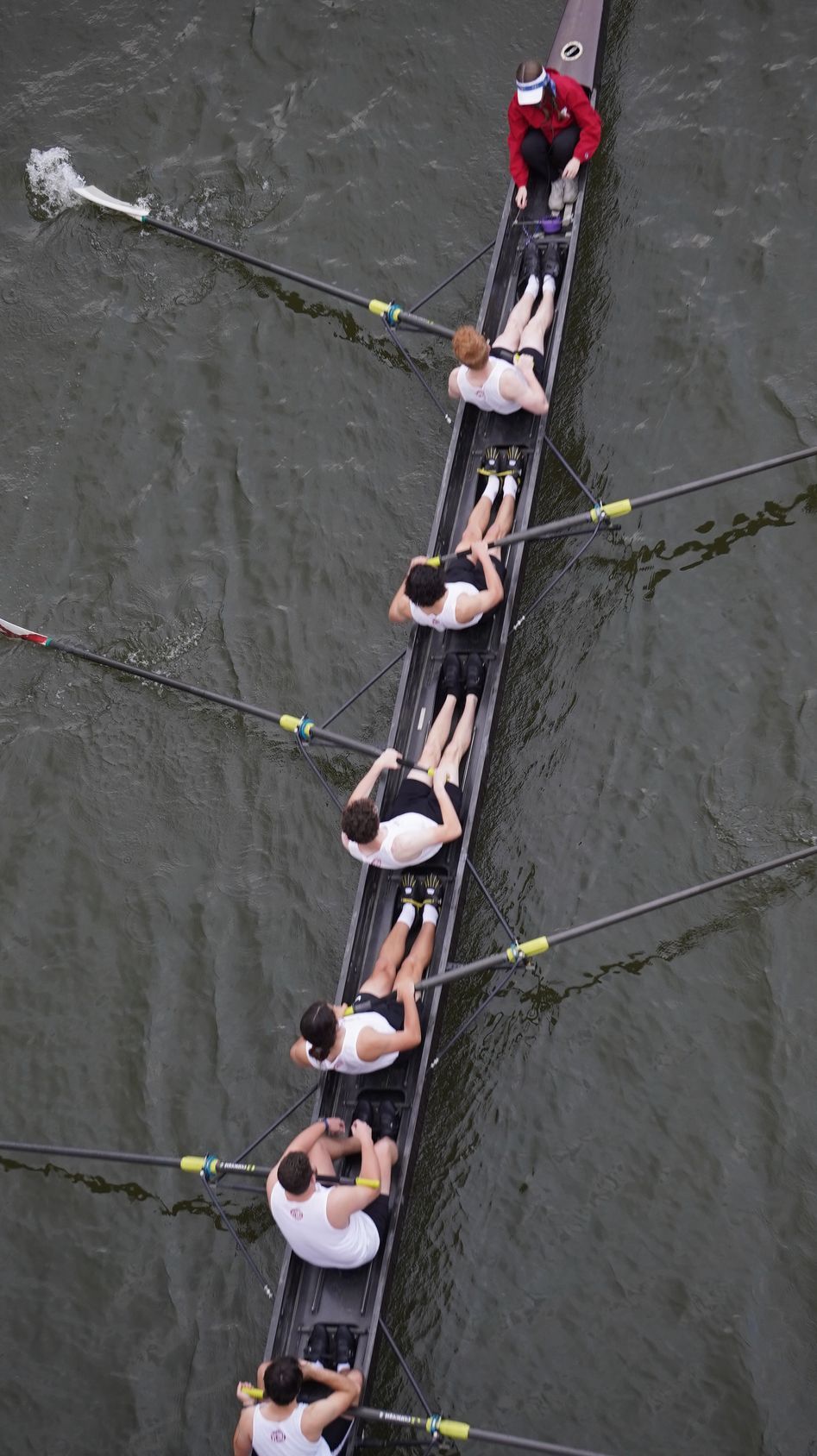 A rowing team in a long boat on water, viewed from above. Team members, in white and black, row in unison.