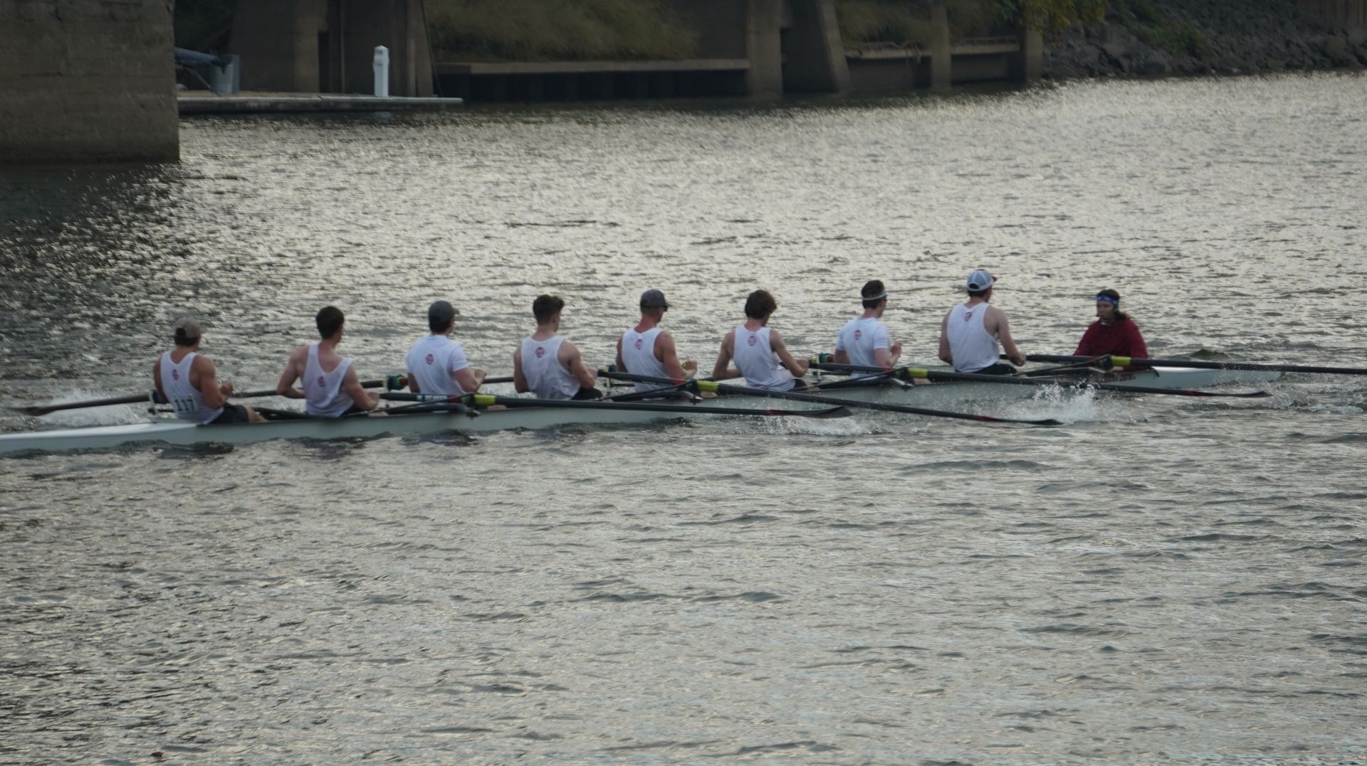 Eight rowers in a boat on a river, backs to the viewer, with water splashing behind them. The setting is outdoors.