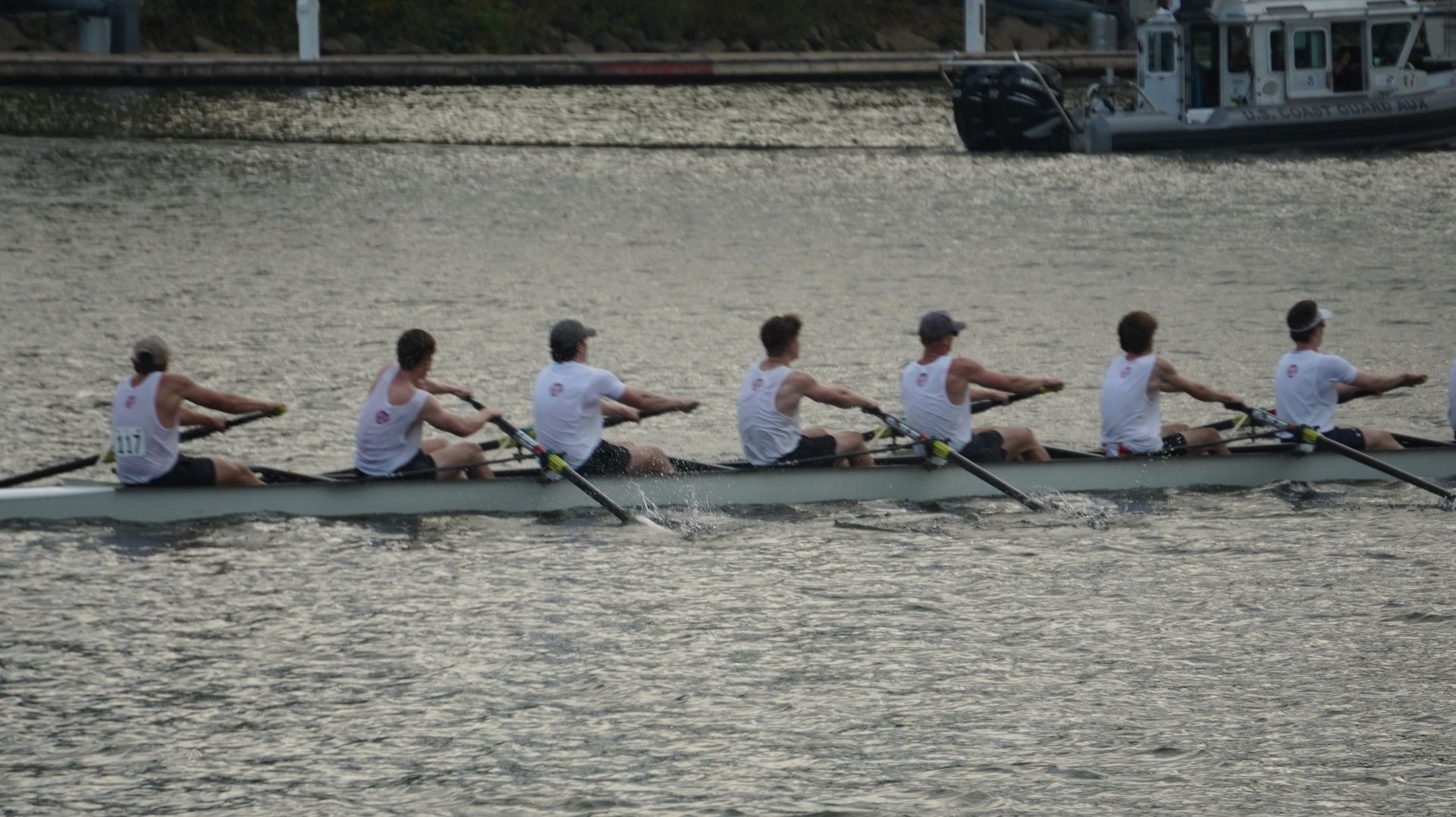 A crew team rowing on a body of water. The rowers wear white shirts and black shorts, performing in unison.