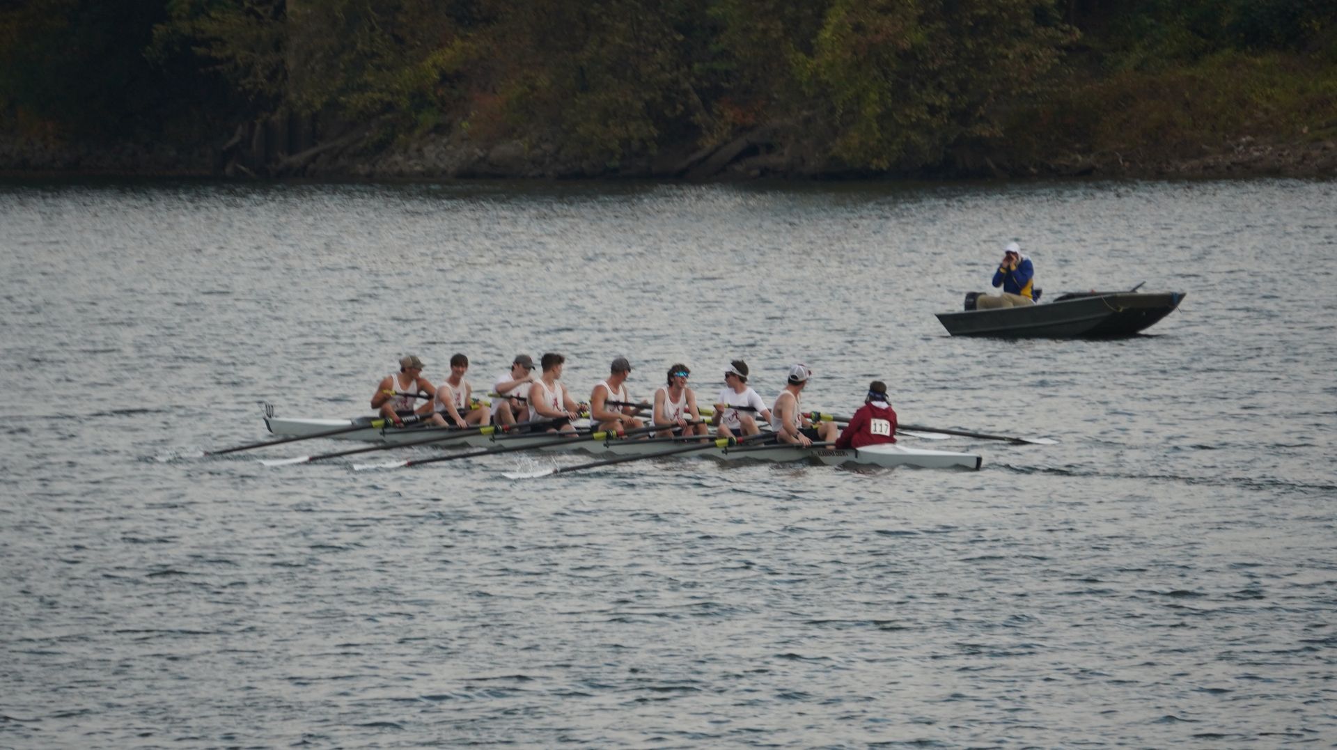 Rowing team on water with oars, accompanied by a motorboat, under an overcast sky.